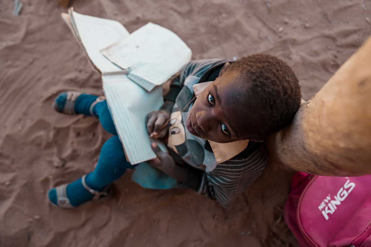 Bambino seduto per terra con un quaderno e una matita in mano che guarda verso la camera.