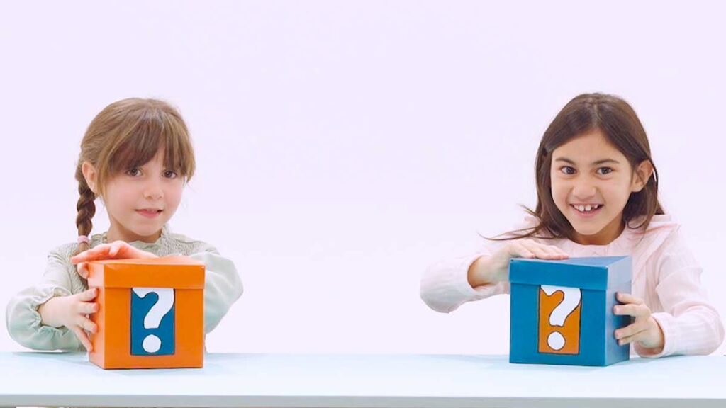 Due bambine sorridenti e incuriosite tengono davabti a loro una scatola chiusa con un punto di domanda scritto sopra.