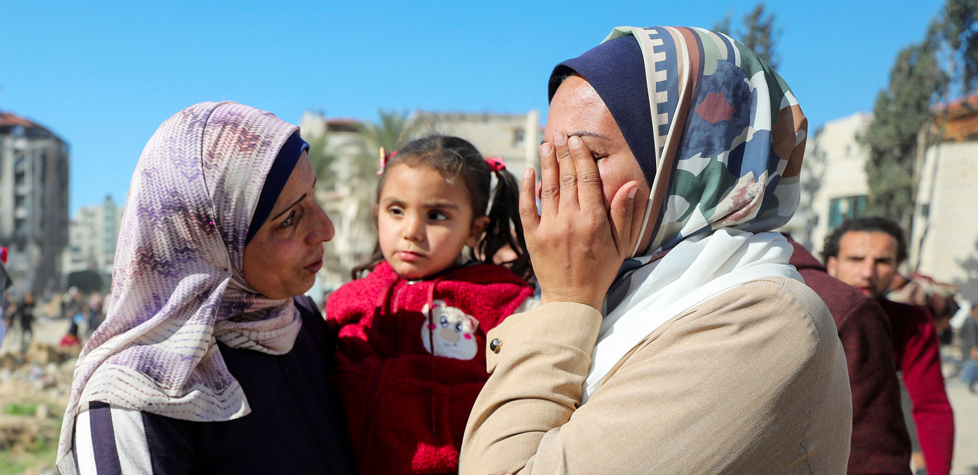 Una donna palestinese si copre il viso con entrambe le mani disperata. Di fronte a lei una donna la guarda con aria triste tenendo n braccio un bambino.