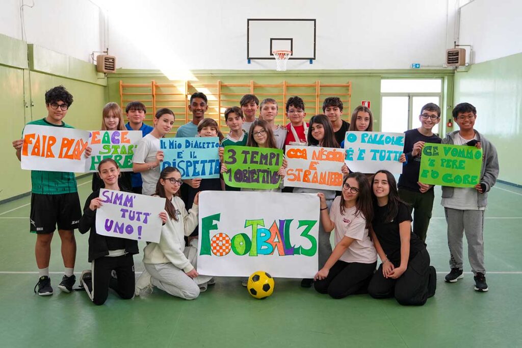 Un gruppo di ragazzi e ragazze in palestra tengono in mano cartelli colorati scritti da loro con Football3