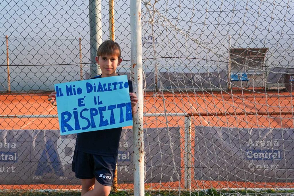 Un ragazzo appoggiato alla porta di una campo da calcio regge un cartello colorato con scritto :il mio dialetto è il rispetto.