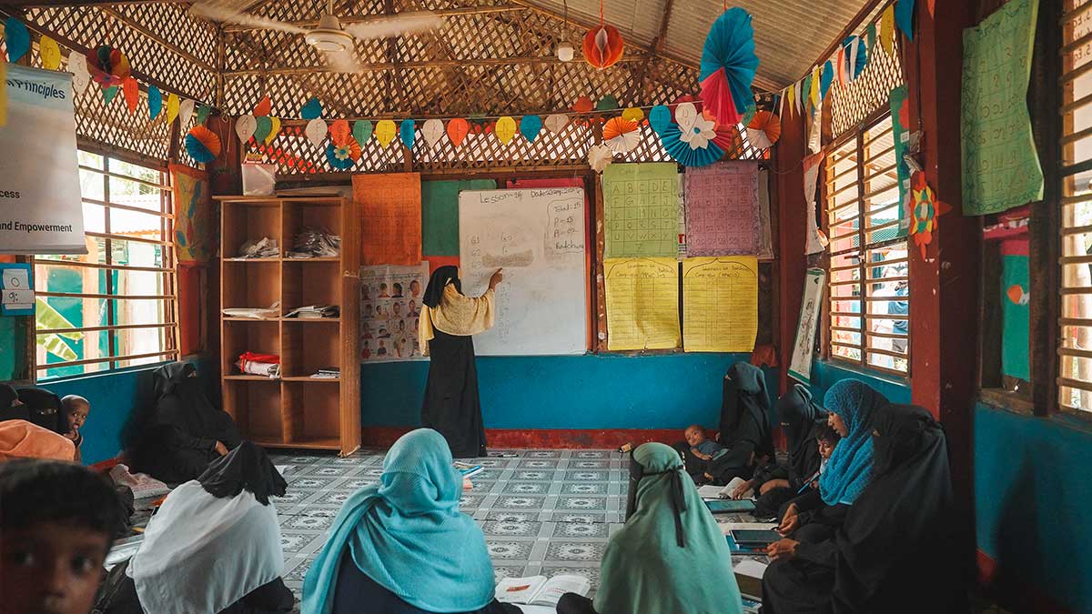 Un'aula adibita a scuola accoglie alcune donne Rohingya sedute a terra, mentre l'insegnante spiega alla lavagna. Ci troviamo in un campo profughi di Cox's Bazar.