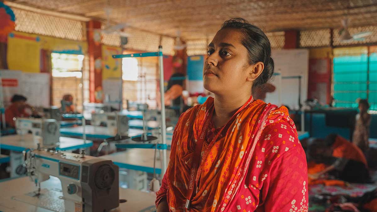 Farzana in primo piano, fa parte dello staff di ActionAid Bangladesh e lavora in uno degli spazi sicuri per donne e ragazze a Cox’s Bazar. Nello sfondo si intravedono postazioni con macchine da cucire.
