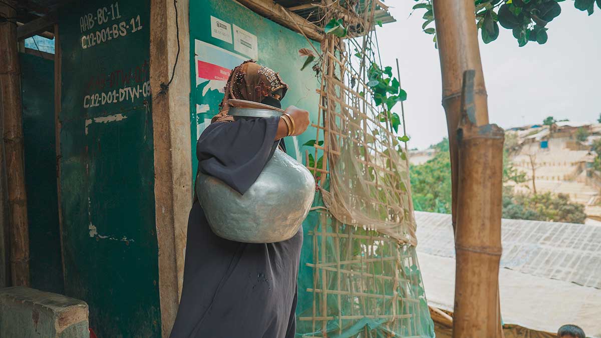 Una signora Rohingya, Esce da un edificio con una grande giara (probabilmente piena di acqua). Ha il velo in testo e il viso coperto. Si trova nel campo di rifugiati di Cox's Bazar