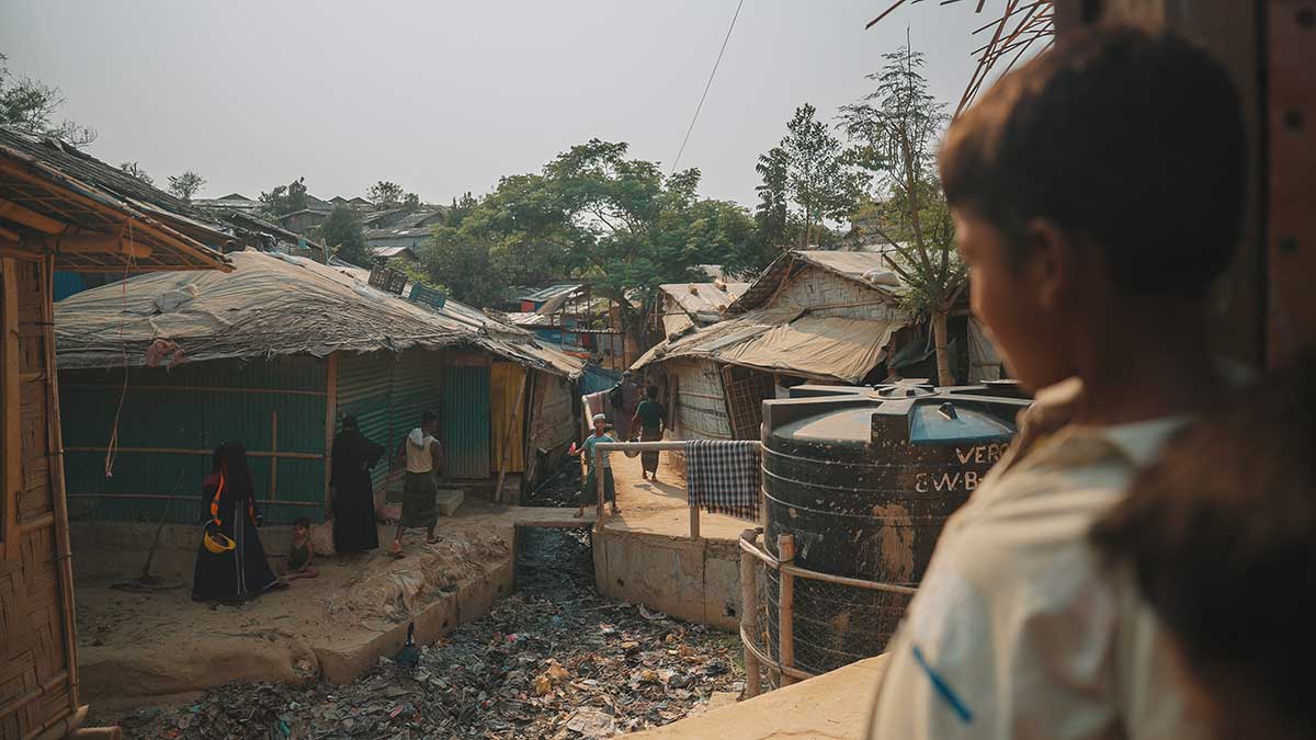 Un bambino Rohingya, gurda fuori da una finesta. Sullo sfondo si intravedono le baracche e alcune persone che vivono nel centro di Cox's Bazar.