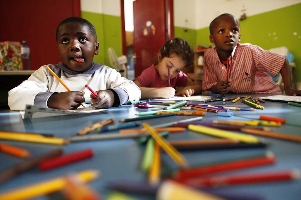 Due bambini di origine africana stanndo disegnando insieme a una loro compagna di scuola italiana tra fogli e tanti pastelli colorati.