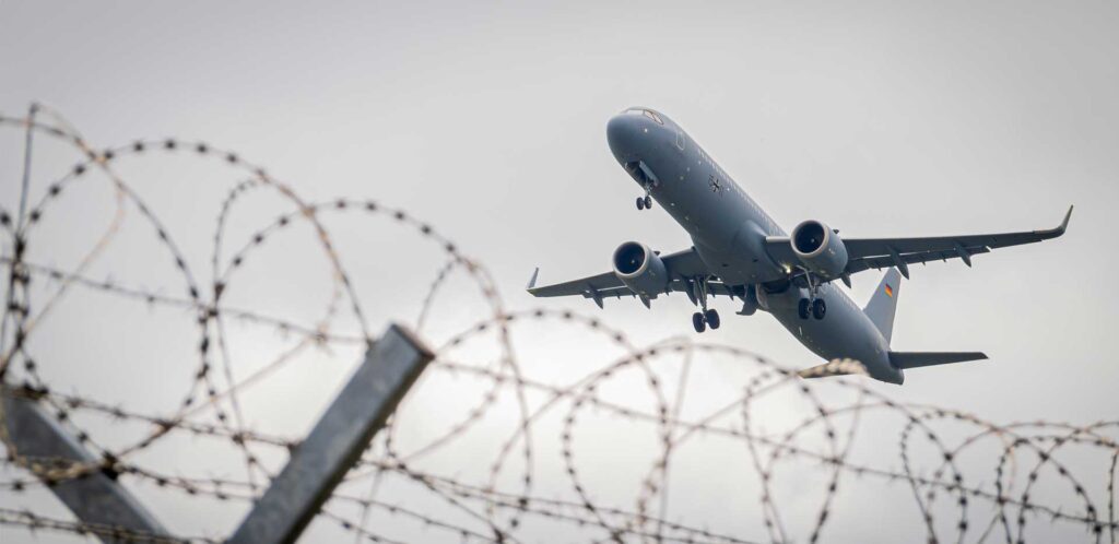Immagine di copertina del report Trattenuti: un aereo in decollo con in primo piano un recinto di filo spinato