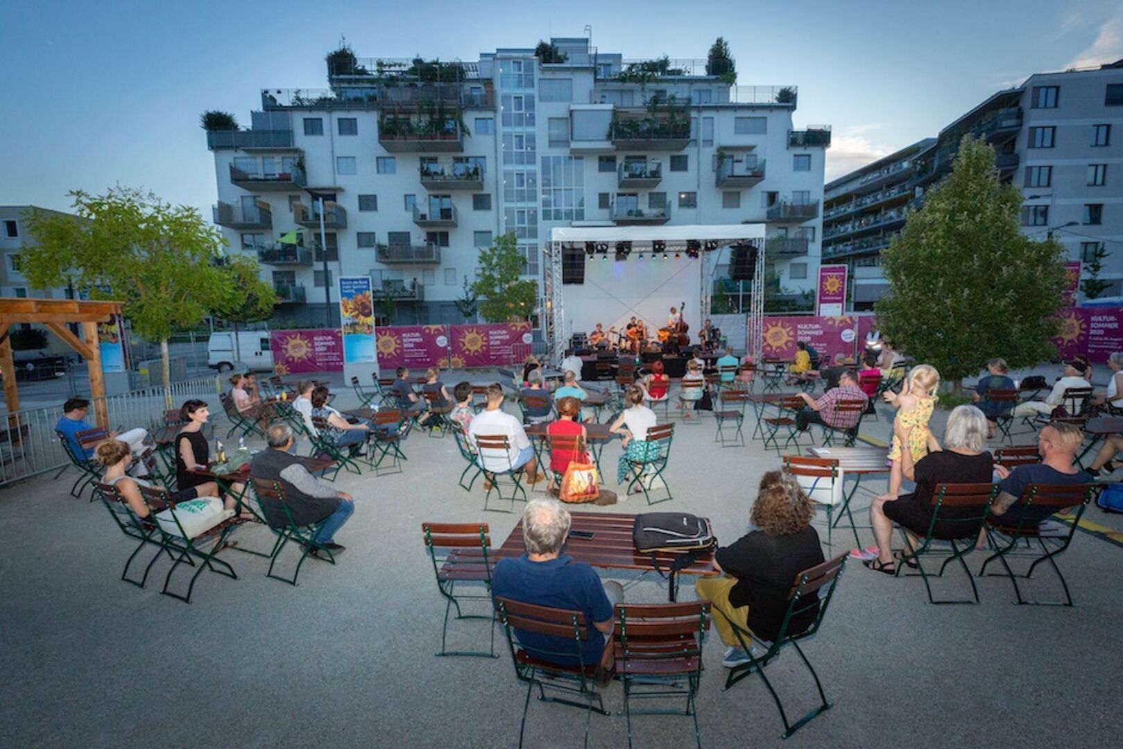 Grüpchen von Menschen sitzend, Holztische, eine Bühne und Band auf einem einem offenen Platz. Häuser im Hintergrund, früher Abend.
