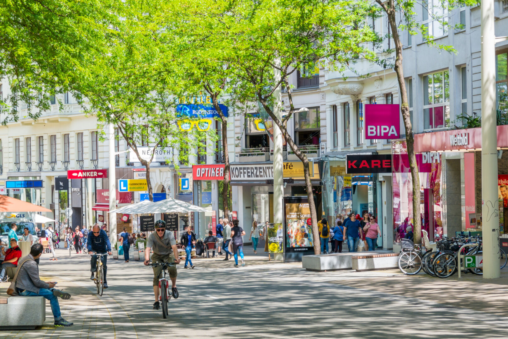 Radfahrer und Passanten auf einer belebten Einkaufsstraße, verschiedene Shops
