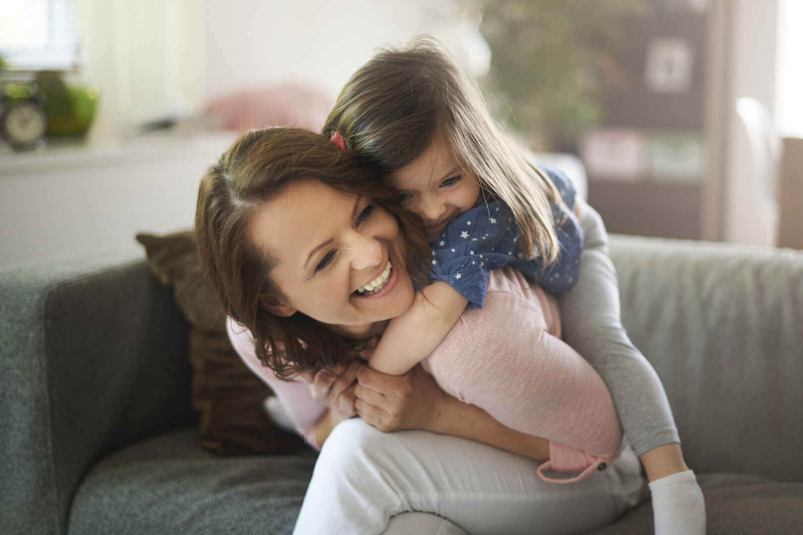 Ein kleines Mädchen umarmt von hinten eine lachende junge Frau | Credit: iStock.com/gpointstudio