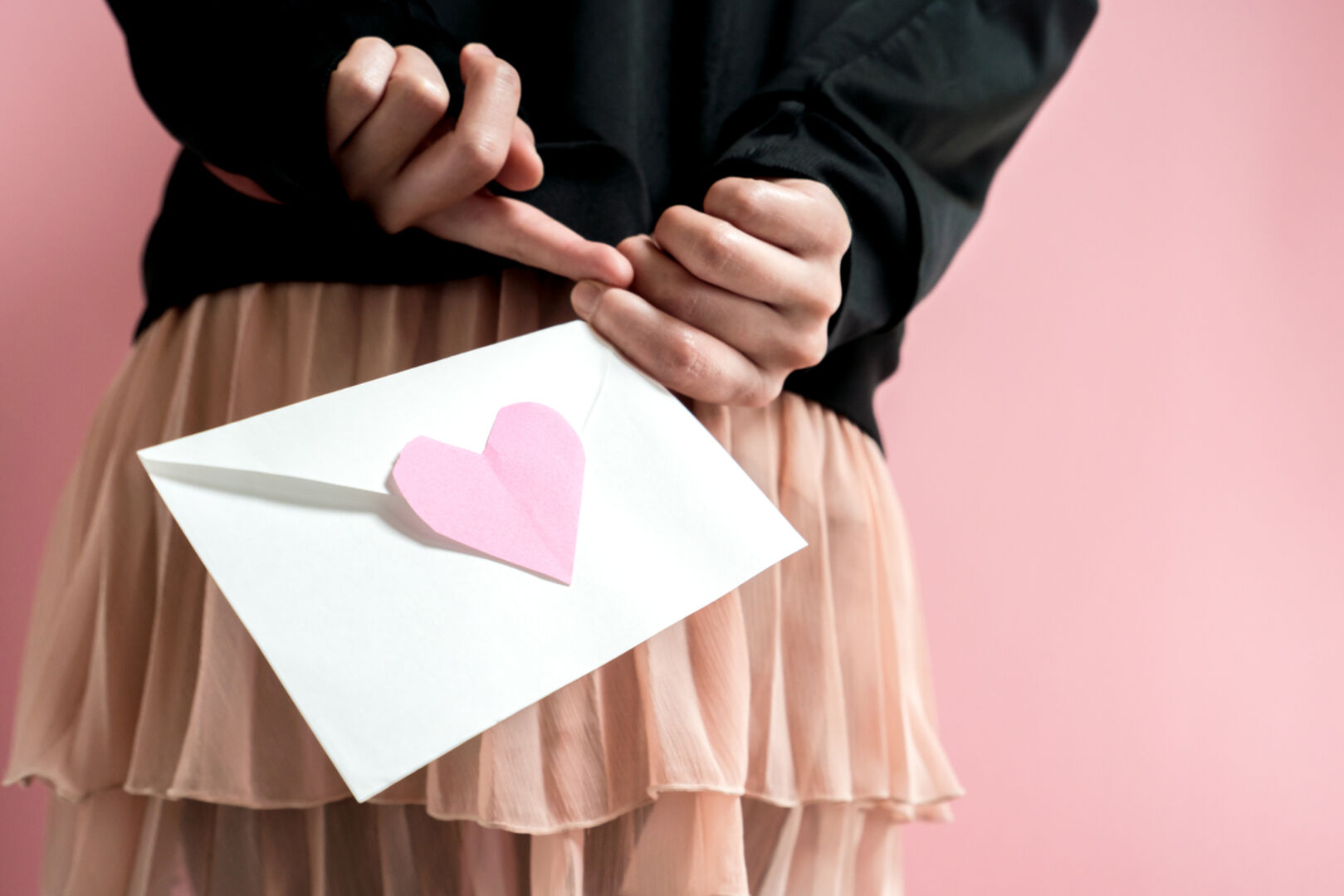 Ein Mädchen hält einen Brief mit einem rosa Herz hinter dem Rücken. Man sieht nur ihre Hände, den Brief und ihren Rock.
