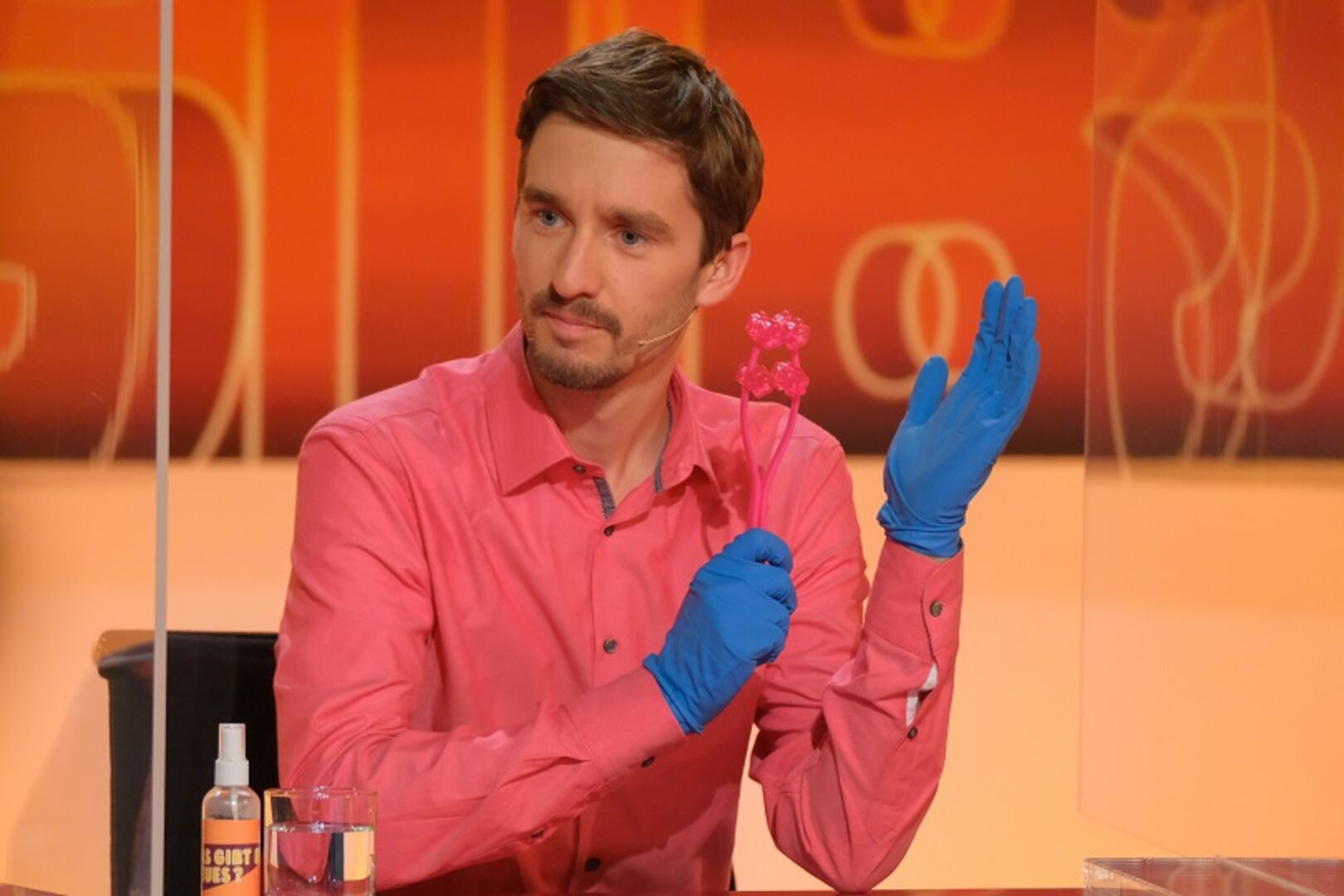 Clemens Maria Schreiner in einem rosa Hemd und blauen Handschuhen in der Sendung "Was gibt es Neues?"