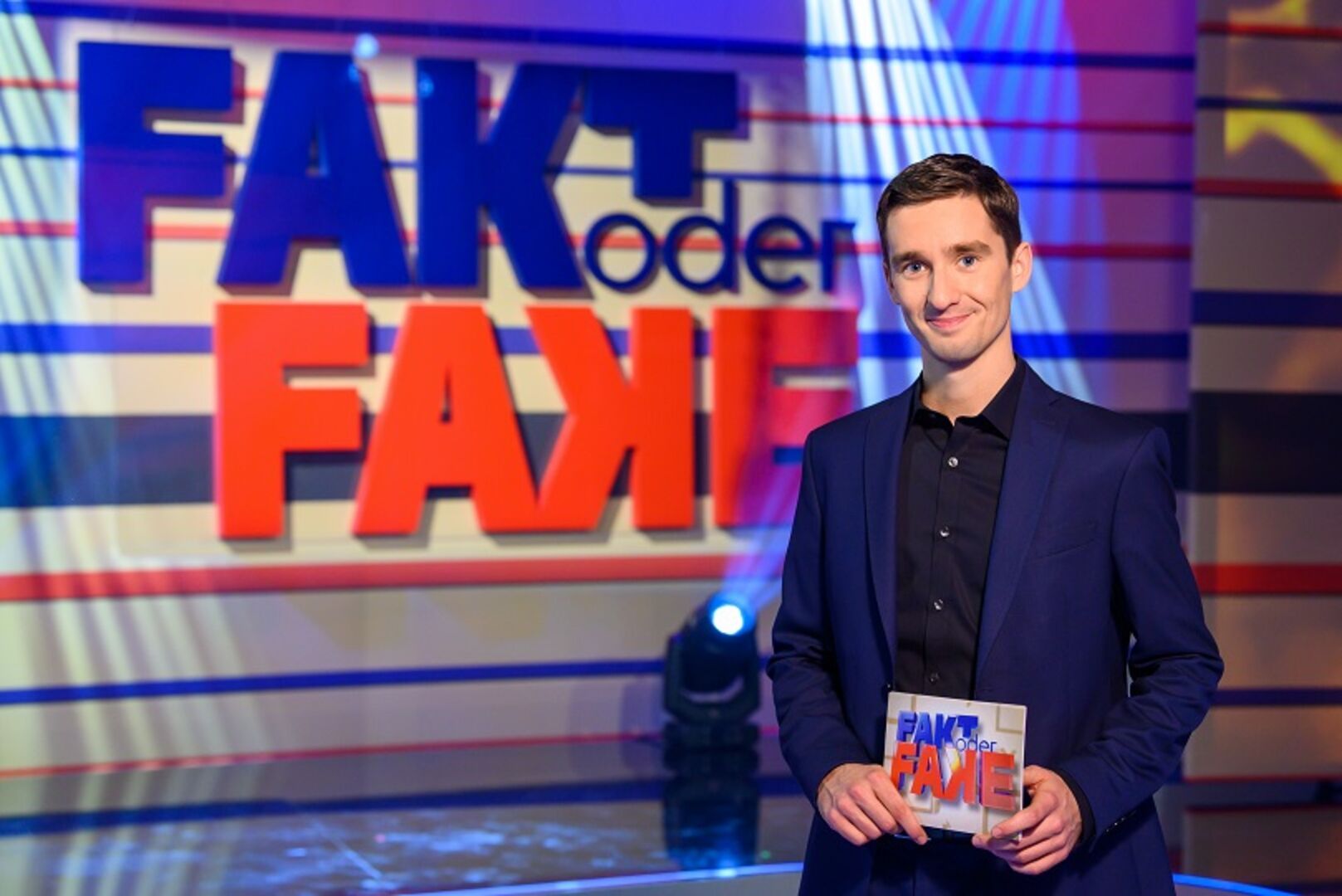 Moderator Clemens Maria Schreiner mit Moderationskarten vor dem Logo der Sendung "Fakt oder Fake".