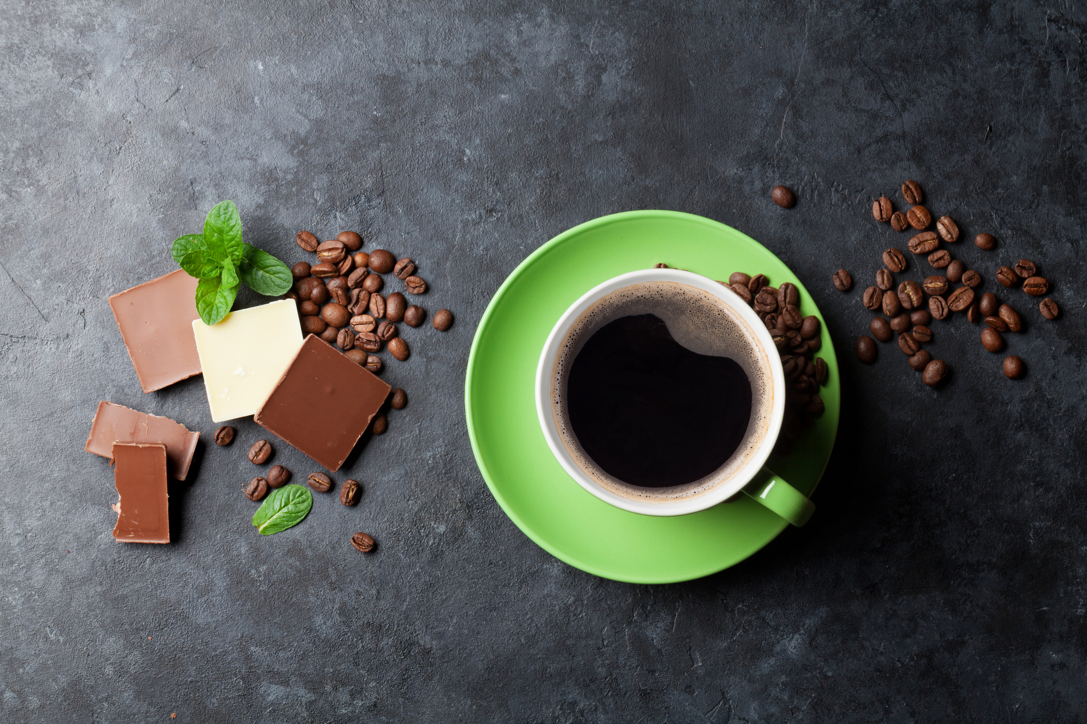Eine grüne Tasse mit schwarzem Kaffee auf einem mit mehreren kleinen Stücken Schokolade und Minzblättern dekorierten Tisch.