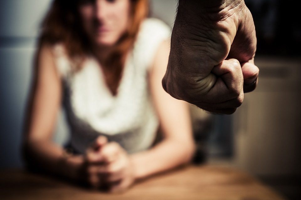 Unscharf im Hintergrund sieht man eine Frau sitzen. Sie hat die Hände gefaltet. Im Vordergrund eine geballte Männerfaust.