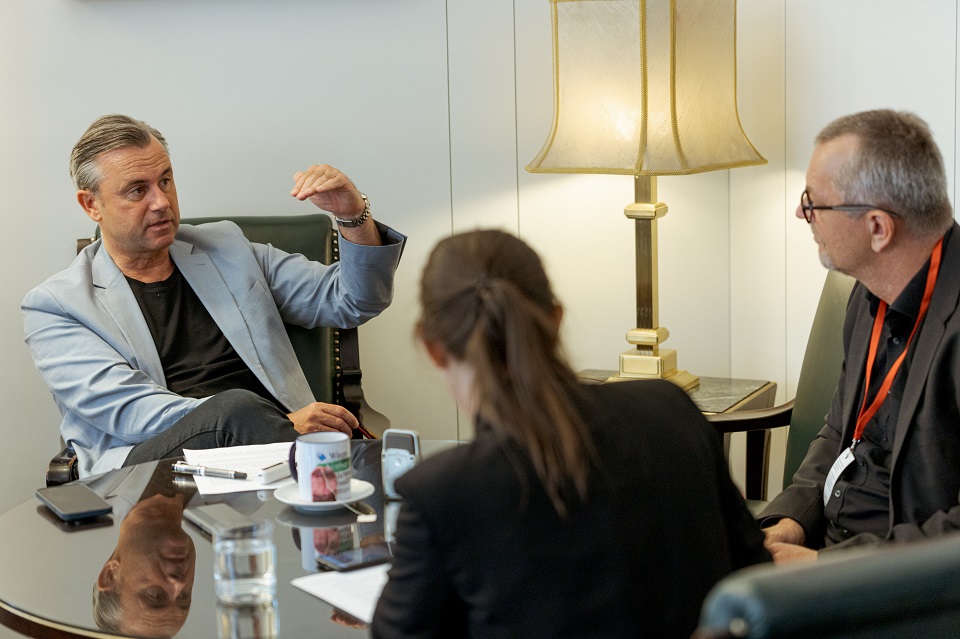 Eine Frau und ein Mann sitzen mit dem Rücken zur Kamera. Norbert Hofer spricht und gestikuliert.
