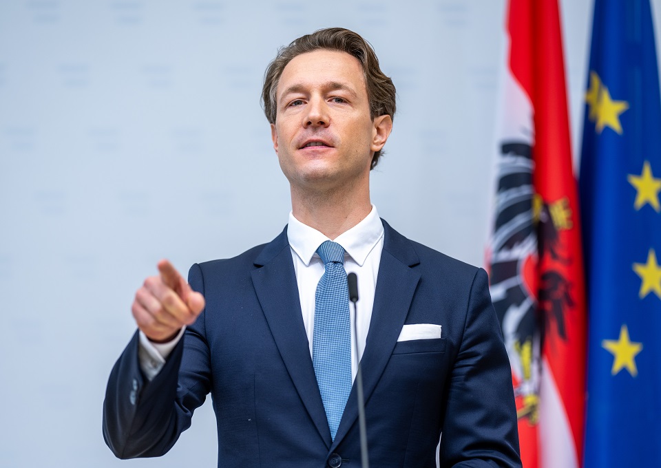 Gernot Blümel steht vor einem Mikrofon, spricht und deutet mit dem Zeigefinger in die Kamera. Hinter ihm österreichische und europäische Flagge.