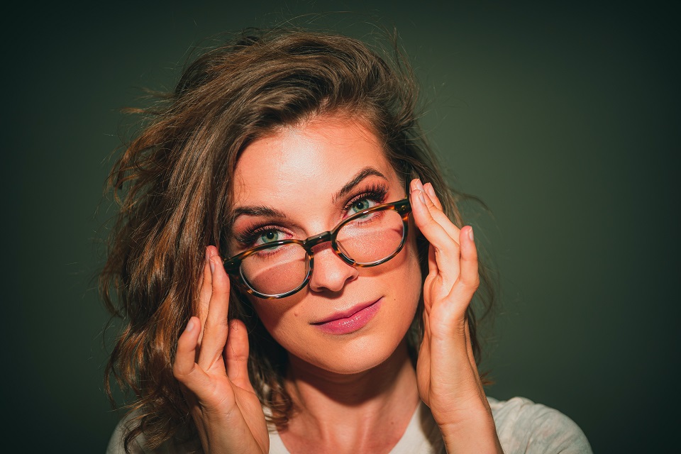 Eine Frau mit langen Haaren greift mit beiden Händen an ihre Brille. Sie schaut über den Rand der Brille und hält den Kopf leicht lief.