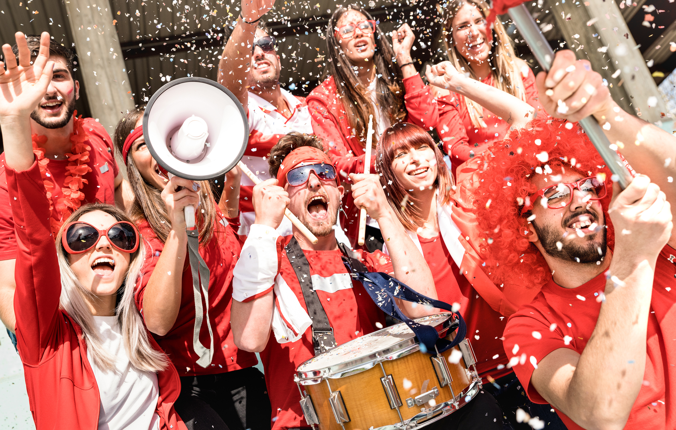 Eine Gruppe von Fußballfans feuert ihr Team mit Instrumenten und einem Megaphon in einem Konfetti-Regen stehend an