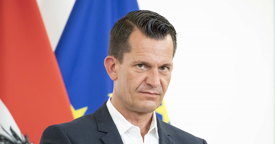 Porträtaufnahme von Gesundheitsminister Wolfgang Mückstein vor einer österreichischen und einer europäischen Flagge