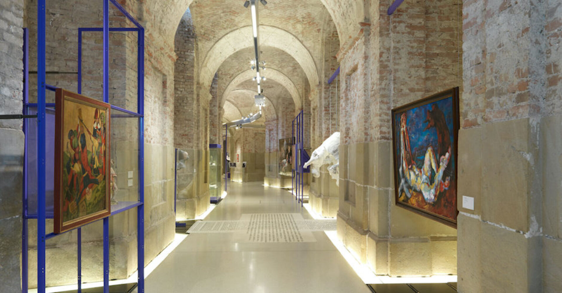 Ein gemauerter, gezielt ausgeleuchteter Gang mit Kunstwerken und Gemälden
