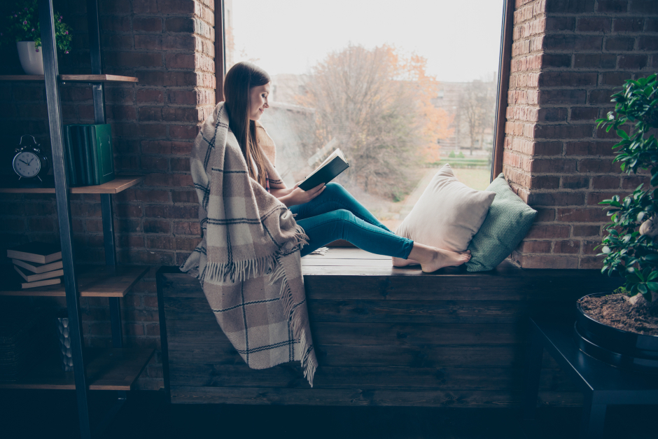 Frau sitzt beim Fenster und liest ein Buch | Credit: iStock.com/Deagreez
