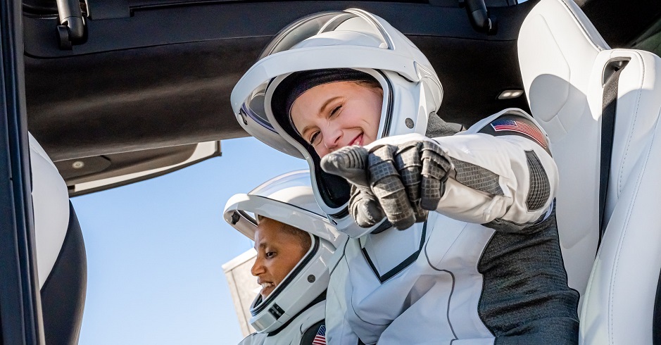 Zwei Frauen in Raumanzügen sitzen gut gelaunt und aufgekratzt in einem Tesla