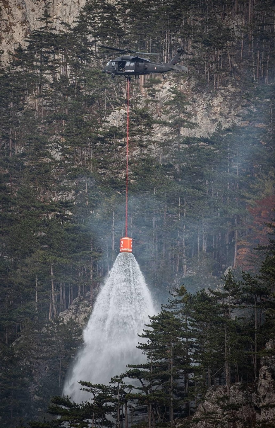 Ein Bundesheer-Hubschrauber unterstützt die Löscharbeiten. Er öffnet einen Löschkübel über dem brennenden Wald.