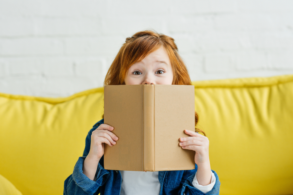 Kleines Mädchen lugt mit den Augen über den Rand ihres Buches und lächelt spitzbübisch in die Kamera | Credit: iStock.com/LightFieldStudios