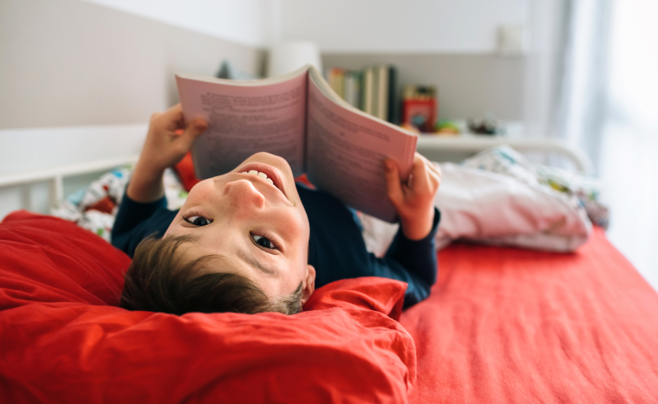 Fröhlicher Junge liegt mit aufgeschlagenem Buch in der Hand auf dem Rücken und lacht über Kopf zurück in die Kamera | Credit: iStock.com/doble-d