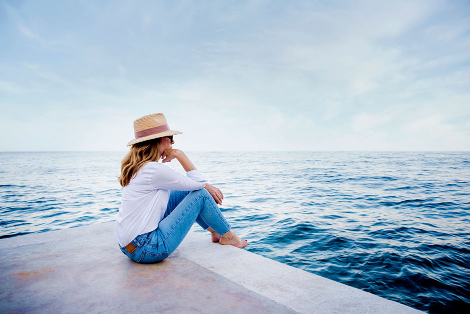 Frau mit Hut und Sonnenbrille blickt auf das offene Meer hinaus | Credit: iStock.com/Szepy