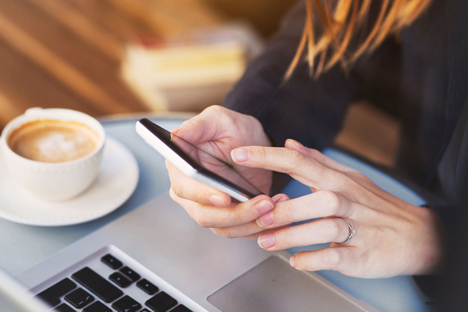 Frau sitzt bei einer Tasse Cappucino vor ihrem Notebook und tippt eine Nachricht auf ihrem Smartphone | Credit: iStock.com/anyaberkut