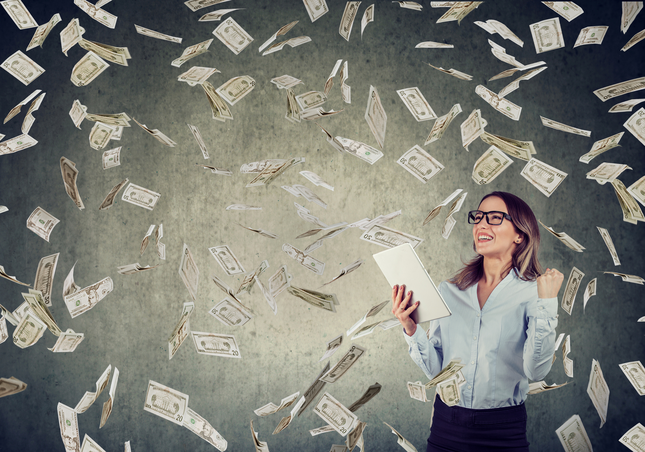 Eine junge Frau im Business-Outfit und mit Tablet in der Hand ballt die Faust unter einem Geldregen zur Jubelgeste