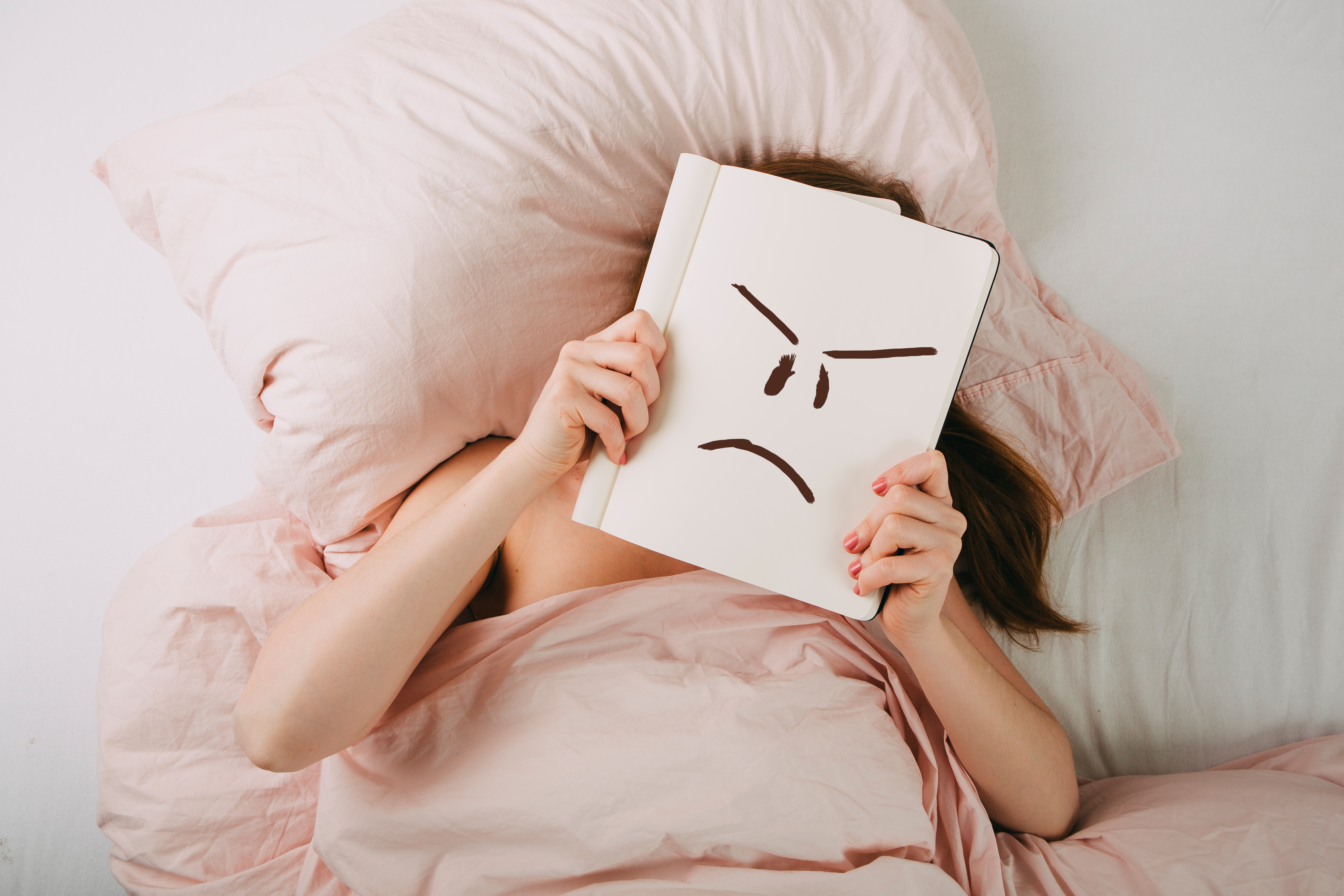 Frau liegt im Bett und hält sich einen Block über das Gesicht