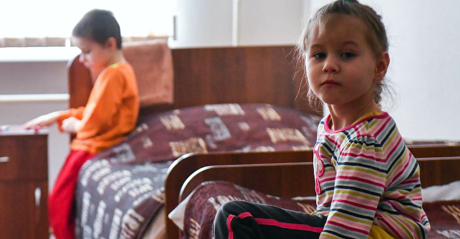 Mädchen sitzt am Bett, im Hintergrund spielt ein Junge
