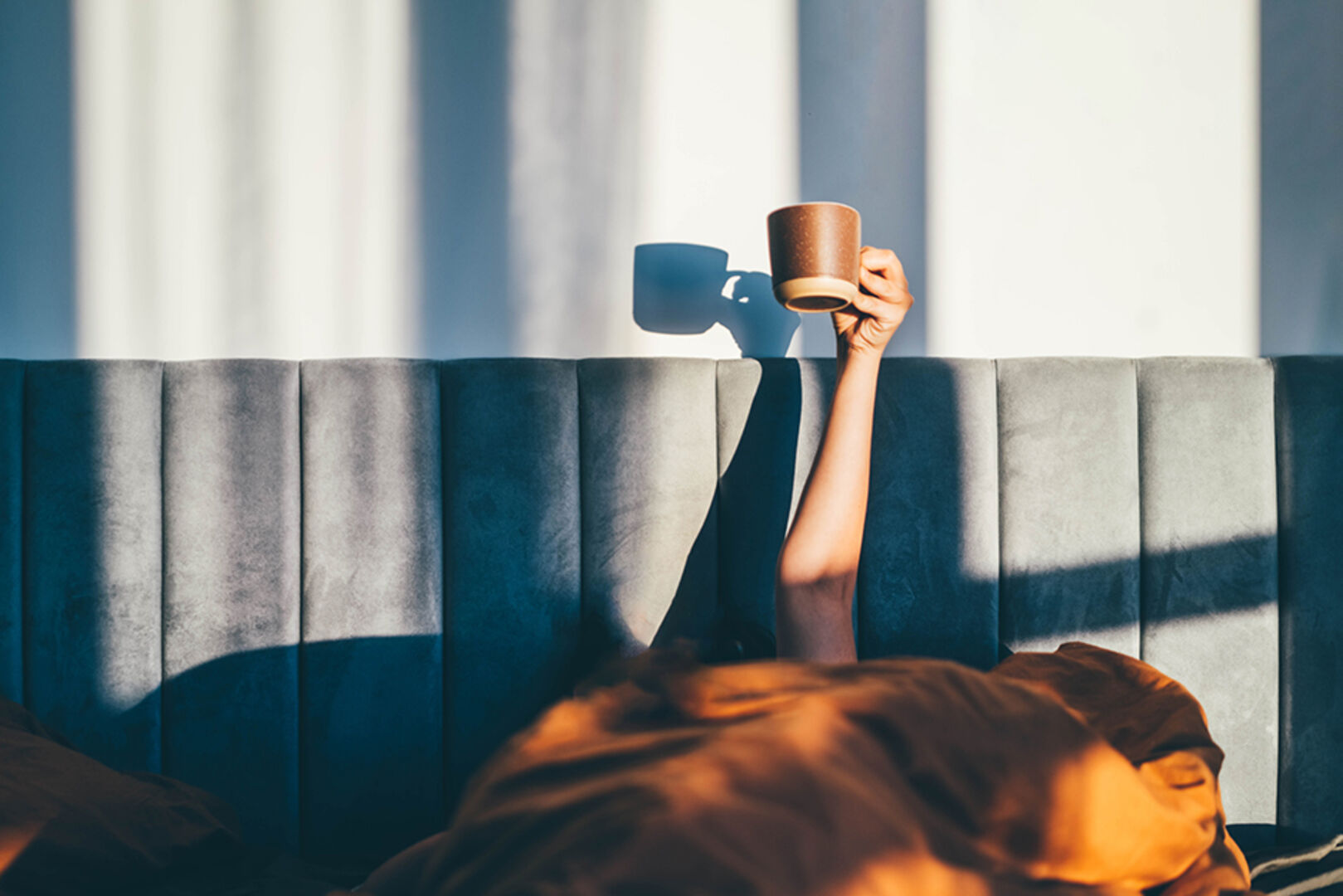 Frau streckt ihren Arm mit einer Tasse in der Hand unter der Bettdecke hervor | Credit: iStock.com/Maria Korneeva