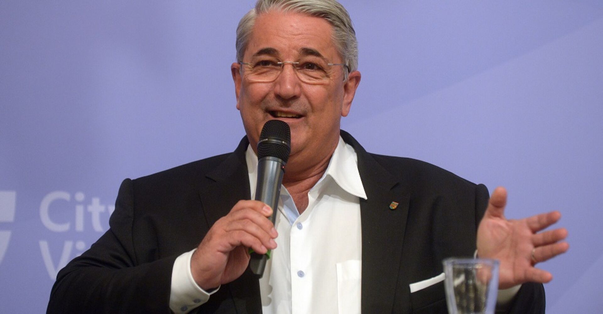 Bezirksvorsteher Ernst Nevrivy vor der Pressewand der Stadt Wien. Er hält ein Mikrofon in der Hand und spricht