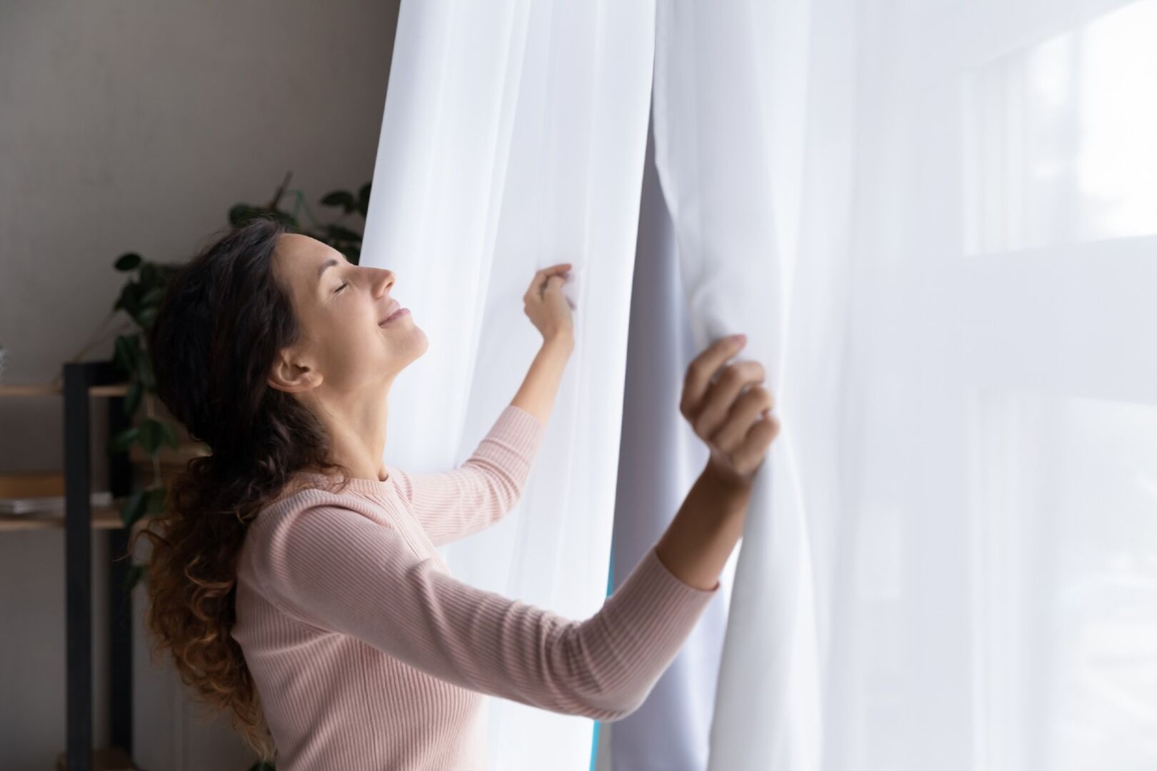 Frau steht vor geöffnetem Fenster mit Vorhängen | Credit: iStock.com/fizkes
