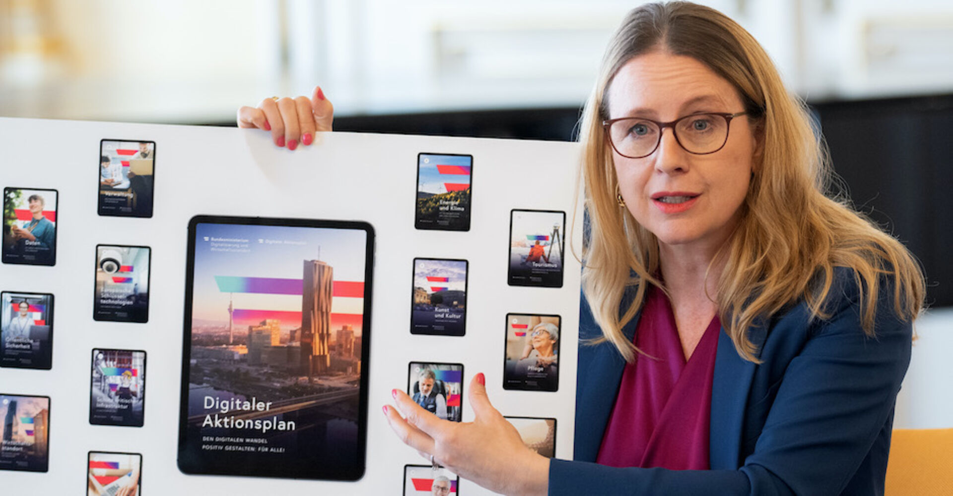 Margarete Schramböck hält einen großformatigen Ausdruck. Darauf zu sehen sind Fotos von Handy- und Tablet-Displays mit dem Slogan "Digitaler Aktionsplan".
