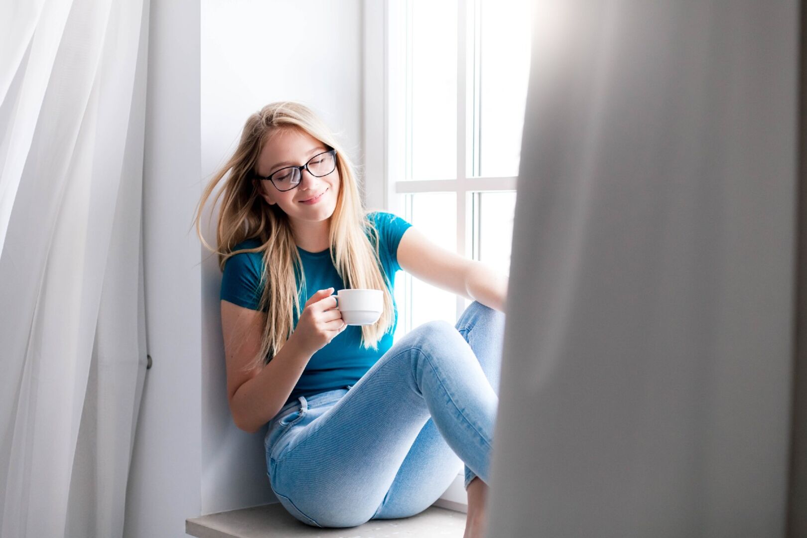 Frau sitzt auf Fensterbank zwischen Vorhänge | Credit: iStock.com/Maryna Andriichenko