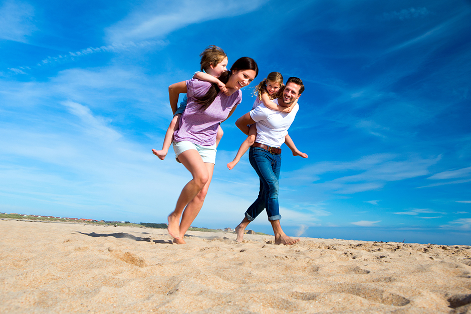 Junges Paar läuft mit seinen beiden Kindern auf dem Rücken fröhlich den Strand entlang | Credit: iStock.com/DGLimages
