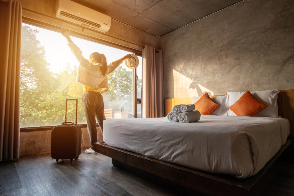 Junge Frau streckt sich am Morgen vor dem Fenster ihres Hotelzimmers durch | Credit: iStock.com/Boyloso