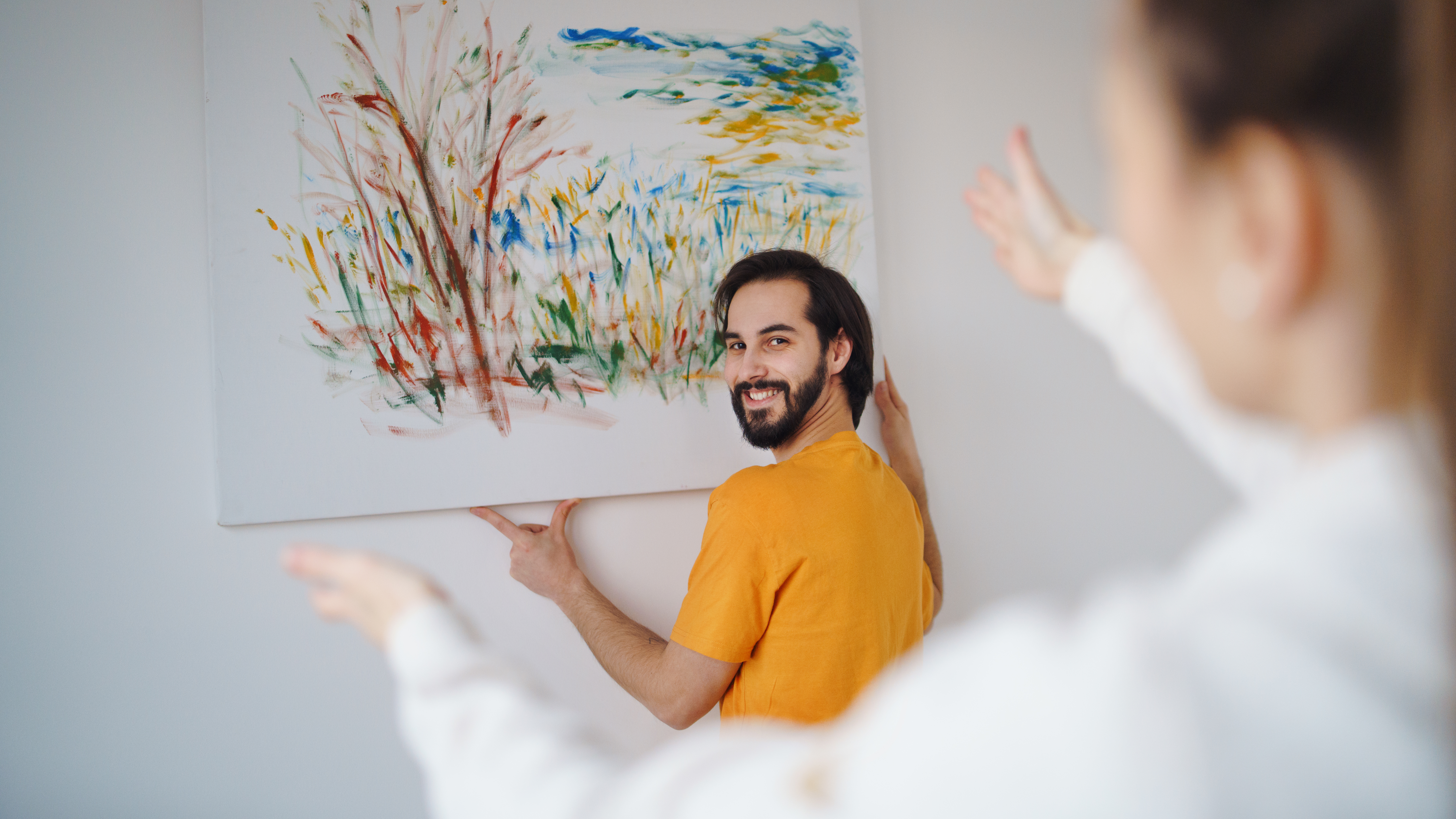 Mann im gelben Shirt hängt ein Bild an die Wand | Credit: iStock.com/Halfpoint
