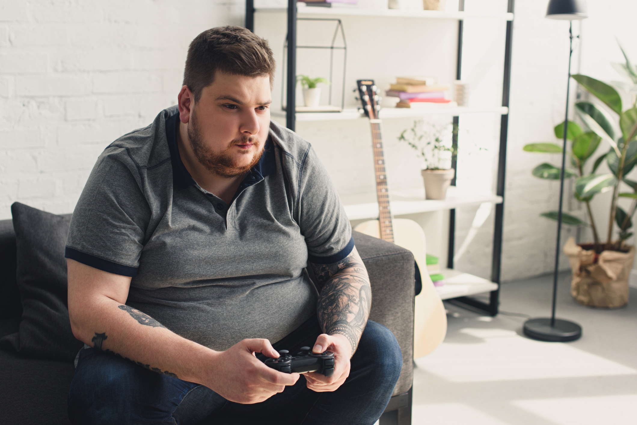 Ein übergewichtiger Mann sitzt mit dem Controller einer Spielkonsole in der Hand auf einer Couch