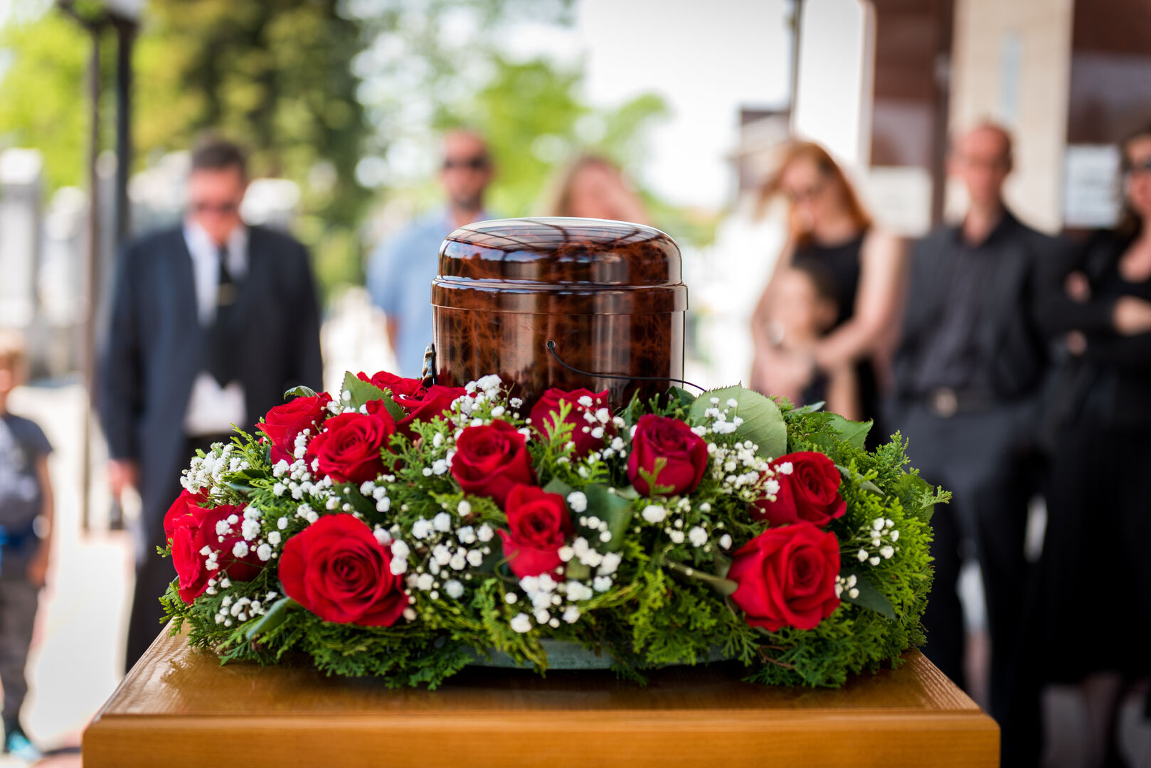 Nahaufnahme einer Urne mit Blumenkranz vor einer sich im Hintergrund befindenden Trauergemeinschaft.