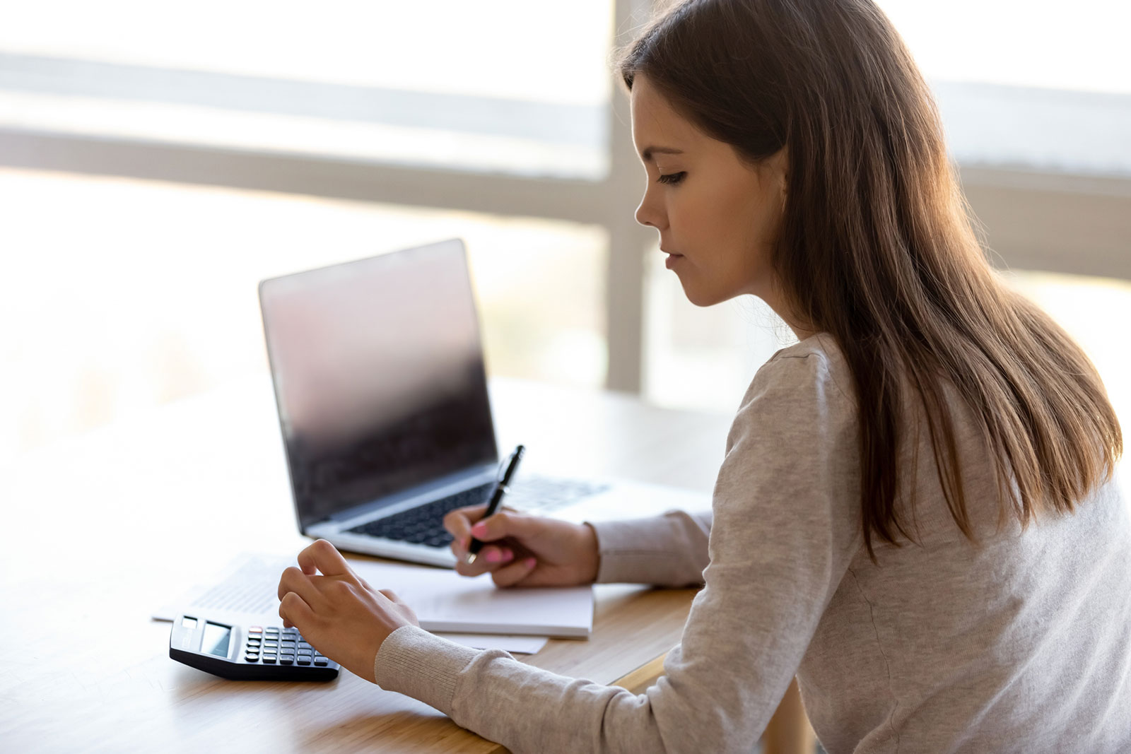 Junge Frau vor ihrem Notebook mit Taschenrechner | Credit: iStock.com/fizkes