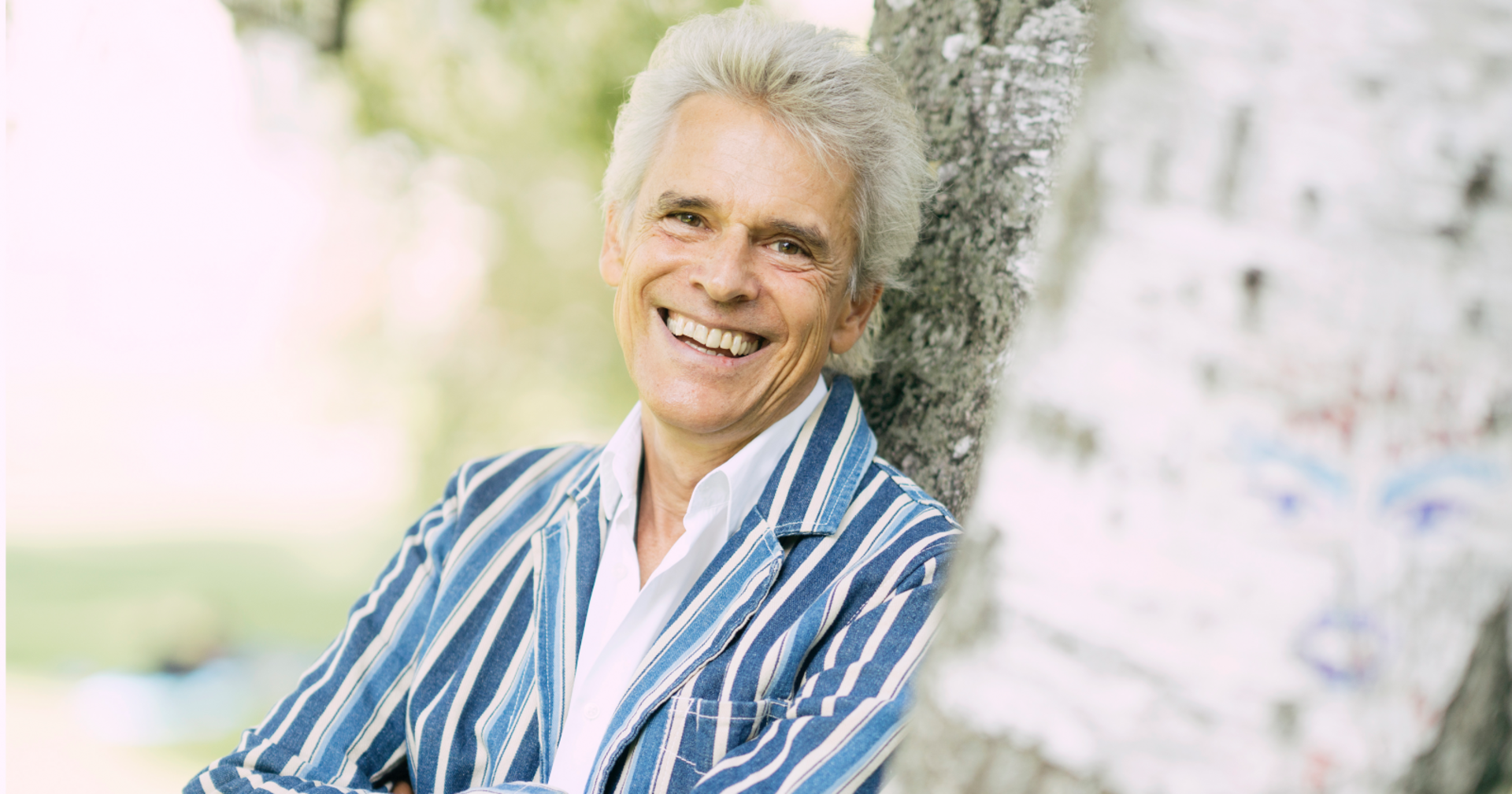 Der in ein blau-weiß-gestreiftes Sakko gekleidete Autor Thomas Brezina, der sich mit verschränkten Armen gegen einen Baum lehnt