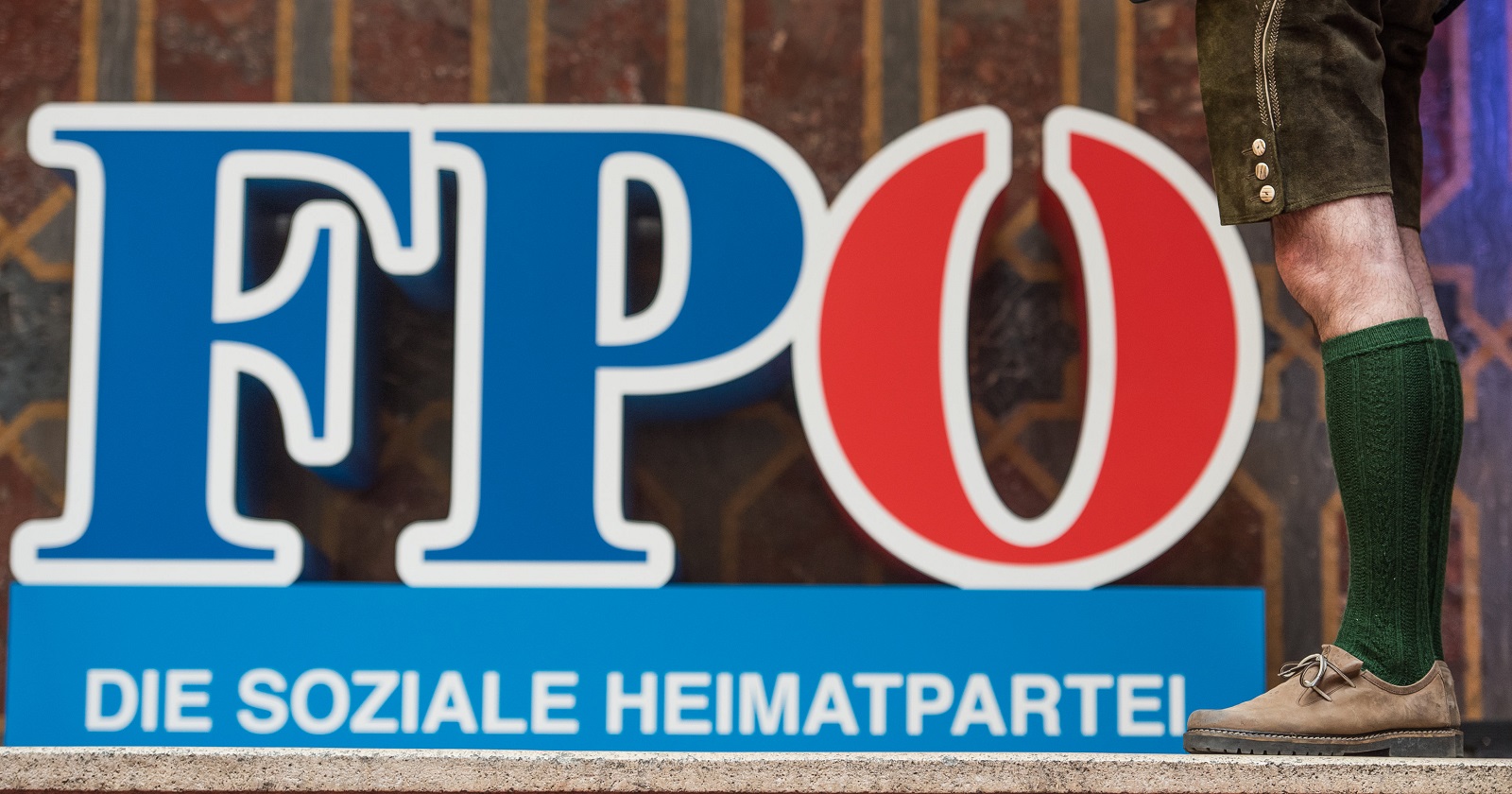 Das Logo der FPÖ mit dem Claim "Die soziale Heimarpartei" als Buchtaben auf einer Bühne stehen. Danebeb in einer Lederhose und Stutzen bekleidete Männerbeine.