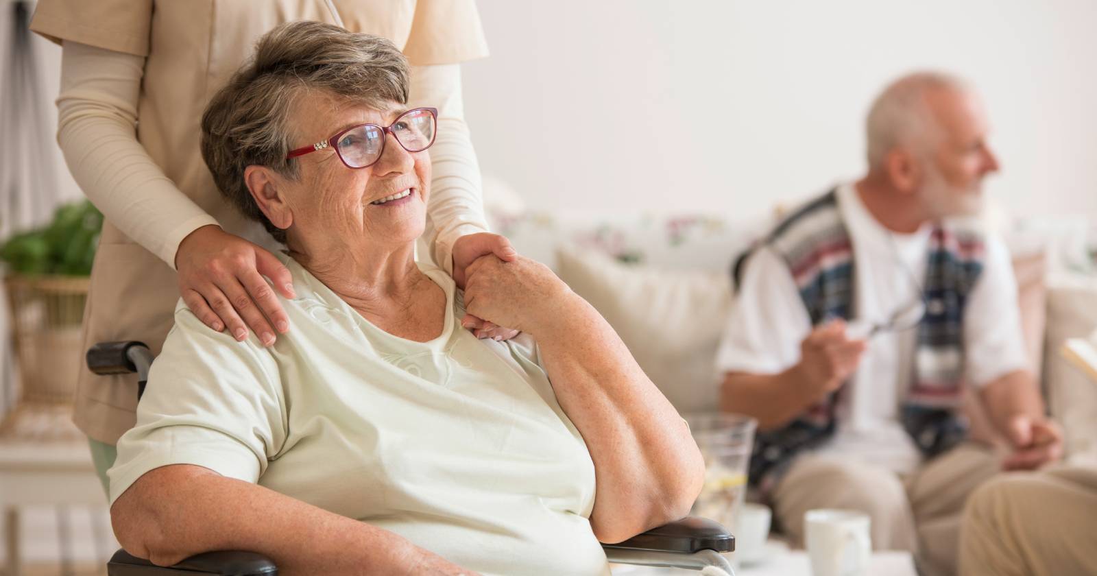 Pflegerin legt ihre Hände auf die Schultern einer älteren Dame im Rollstuhl | Credit: Adobe Stock/Photographee.eu