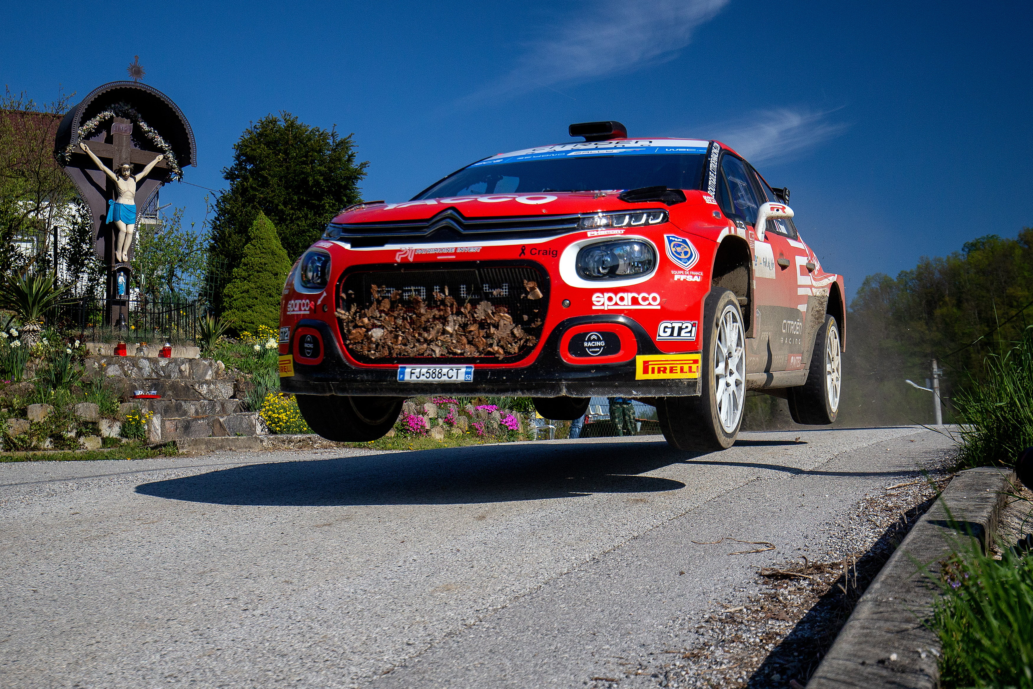 Ein rotes Rallye-Auto, das über eine Straßenkuppe springt - im Hintergrund ist ein Bildstock mit der Darstellung des gekreuzigten Jesus zu sehen